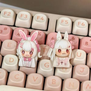 May include: Two custom keycaps featuring anime-style characters with bunny ears, set on a keyboard. The keycaps are white and pink, with detailed facial features and clothing. The keyboard keys are a mix of white and pink.