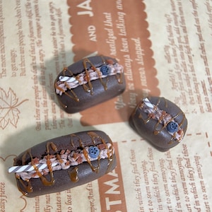 Può includere: Tre pasticcini in miniatura ripieni di cioccolato e caramello con un mirtillo e una cannuccia in cima. I pasticcini sono su una superficie di carta marrone.