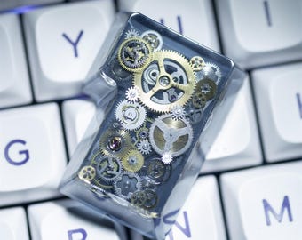 May include: A custom mechanical keyboard keycap featuring a steampunk design. The keycap is clear with a dark background, filled with various sizes of gold and silver gears. The keycap is resting on a white keyboard.
