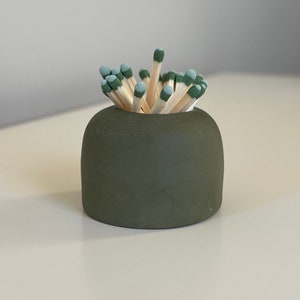 May include: A small, olive green ceramic match holder filled with wooden matches. The match heads are a muted teal color. The holder has a rounded shape and sits on a white surface.