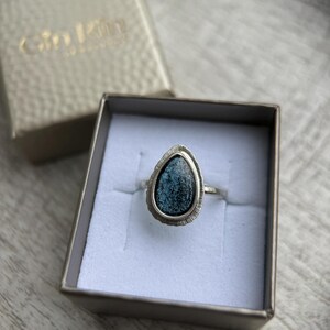 May include: A silver ring featuring a teardrop-shaped blue stone set in a silver bezel. The ring is displayed in a white box with a gold label that reads "Gilded" in cursive.