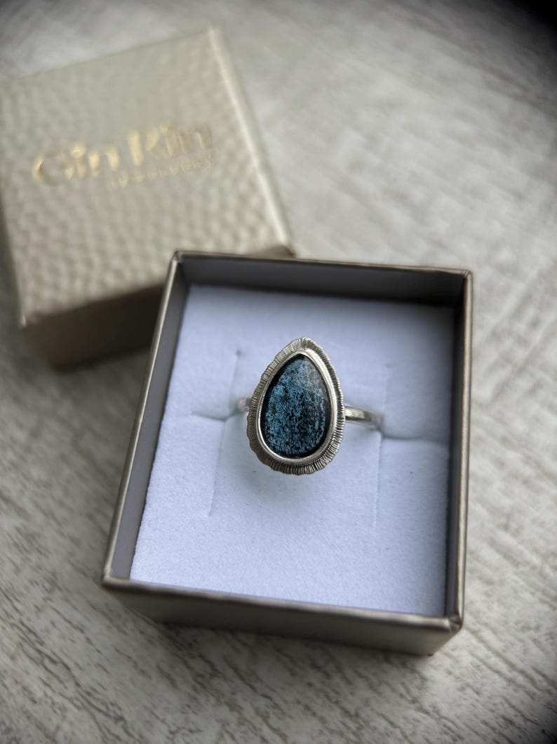 May include: A silver ring featuring a teardrop-shaped blue stone set in a silver bezel. The ring is displayed in a white box with a gold label that reads "Gilded" in cursive.