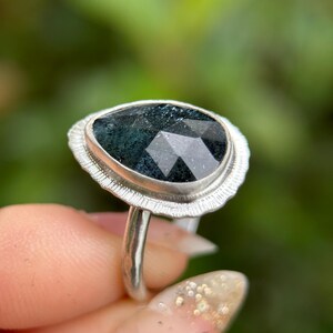 May include: A silver ring featuring a dark green faceted gemstone set within a textured silver bezel. The ring is worn on a finger.