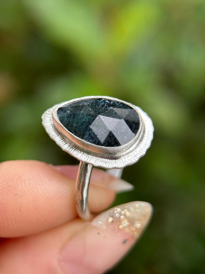 May include: A silver ring featuring a dark green faceted gemstone set within a textured silver bezel. The ring is worn on a finger.