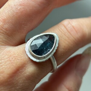 May include: A silver ring featuring a dark blue gemstone set in a textured silver bezel. The ring is worn on a finger.