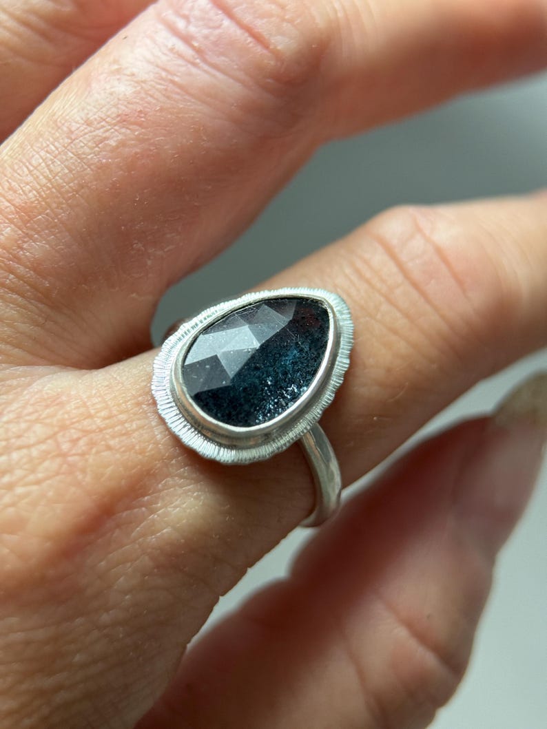 May include: A silver ring featuring a dark blue gemstone set in a textured silver bezel. The ring is worn on a finger.