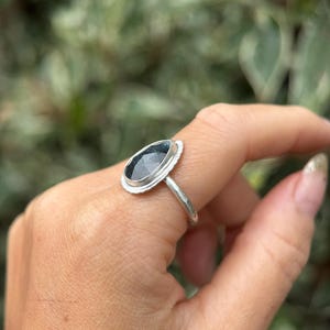 May include: A silver ring with a dark blue gemstone set in a bezel setting. The ring is on a finger.