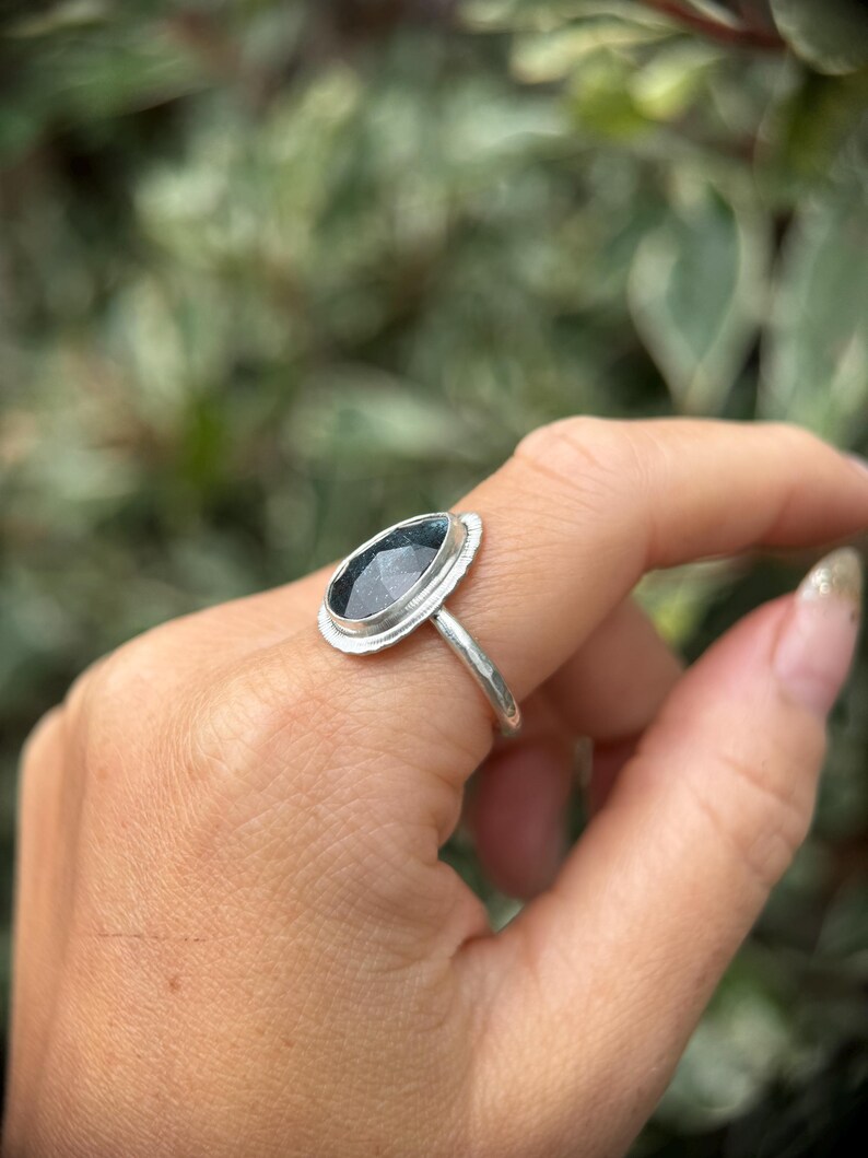 May include: A silver ring with a dark blue gemstone set in a bezel setting. The ring is on a finger.