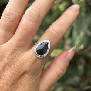 May include: A silver ring featuring a black pear-shaped gemstone set in a bezel setting. The ring is worn on a finger.