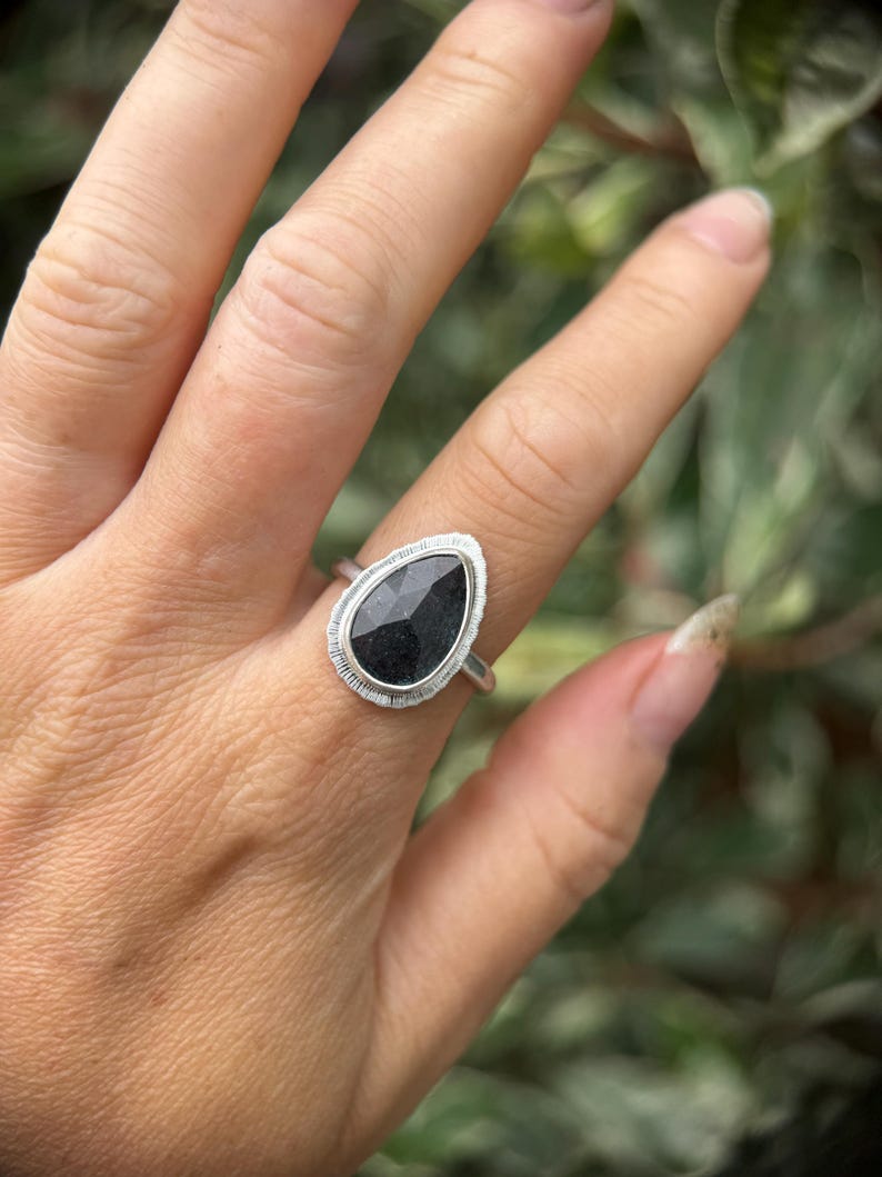 May include: A silver ring featuring a black pear-shaped gemstone set in a bezel setting. The ring is worn on a finger.