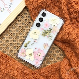 Puede incluir: Una funda transparente para teléfono con flores secas prensadas en su interior. Las flores son blancas, rosas y verdes. La funda está diseñada para un teléfono inteligente con una lente de cámara triple.