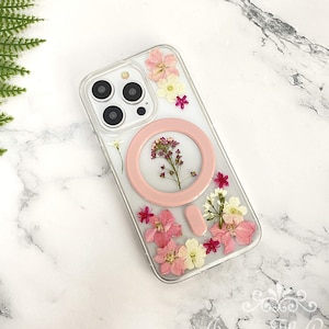 Puede incluir: Una funda de teléfono transparente con un soporte de anillo rosa y flores prensadas. Las flores son rosas, blancas y amarillas.