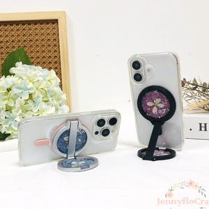 May include: Two clear phone cases with a black and silver phone ring holder. The phone ring holder has a floral design with a purple flower and gold glitter. The phone cases are on a white surface.