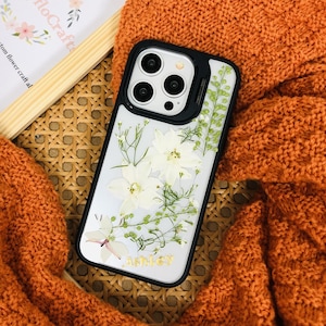 以下が含まれることがあります： 黒い縁取りのある透明な携帯電話ケースで、白い花と緑の葉の入った花柄のデザインです。ケースには「Ashley」という金色の文字が刻まれています。