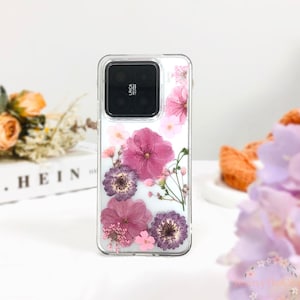Può includere: Una custodia per telefono trasparente con un design floreale. La custodia presenta fiori rosa e viola pressati, tra cui un grande fiore rosa al centro. La custodia è progettata per un telefono con un obiettivo della fotocamera quadrato.