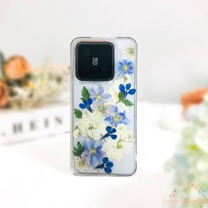 Puede incluir: Una funda transparente para teléfono con un diseño de flores prensadas en azul y blanco. La funda tiene un recorte para el objetivo de la cámara, que está etiquetado como "Leica".