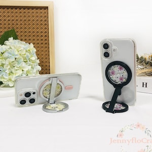 Puede incluir: Dos fundas transparentes para teléfono con soportes de anillo florales. Una funda está tumbada, la otra está de pie. Los soportes de anillo tienen un marco negro y un diseño floral. Las fundas están sobre una superficie blanca.