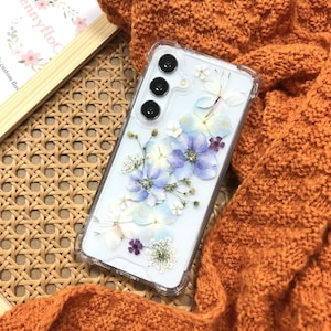 May include: A clear phone case with a pressed flower design. The flowers are blue, white, and purple. The case is designed for a phone with a triple camera lens.