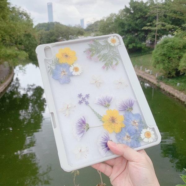 Puede incluir: Una funda transparente para tableta con marco gris claro que presenta un arreglo de flores secas prensadas en tonos amarillo, azul, morado y blanco. El diseño floral incluye margaritas, pétalos pequeños y follaje verde en la parte trasera.