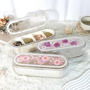 May include: Clear, oval-shaped jewelry boxes with ribbed sides and clear lids. One box is open, revealing compartments with jewelry and makeup sponges. Two other boxes feature floral arrangements under the lids. The boxes are on a fluffy white surface.