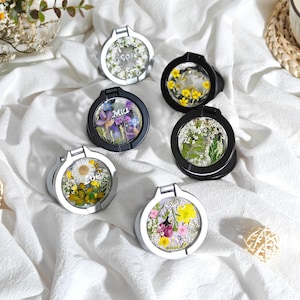 Op de afbeelding: Een verzameling telefoonhouders met bloemmotieven. De ronde houders hebben een transparant midden, met geperste bloemen en de namen "Rose", "Mia" en "Love". De frames zijn zilver en zwart, liggend op een witte stof.