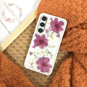 以下が含まれることがあります： クリアな背面と押し花でできたピンクと白の花のデザインが施された白い携帯電話ケース。ケースにはトリプルカメラレンズと背面に「Zoe」という名前が書かれています。