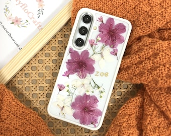 May include: White phone case with a clear back and a design of pressed pink and white flowers. The case has a triple camera lens and the name "Zoe" is written on the back.