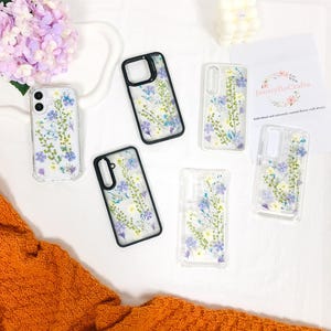 以下が含まれることがあります： 押し花を使った透明な携帯電話ケースの詰め合わせ。ケースには、紫、青、白の色合いのさまざまな花のアレンジメントが施されています。ケースはさまざまな携帯電話モデルに適合するように設計されています。JennyFloCrafts.