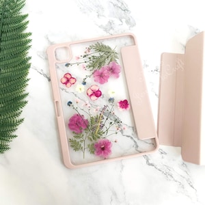 Puede incluir: Una funda para tableta de color rosa claro con un panel trasero transparente que contiene flores prensadas en tonos rosa, morado y blanco. La funda incluye una tapa plegable rosa a juego y un recorte para la lente de la cámara.