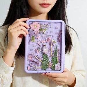 May include: A rectangular lavender-colored case featuring a clear front panel decorated with an arrangement of pressed pink, purple, and white flowers, green ferns, and foliage. The word "June" is written in silver script in the center.
