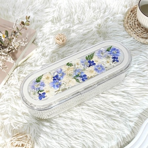 Puede incluir: Un recipiente rectangular transparente con un borde blanco, lleno de flores azules y blancas secas y follaje verde. El recipiente se encuentra sobre una superficie blanca y esponjosa, con un libro y una bola decorativa en el fondo.