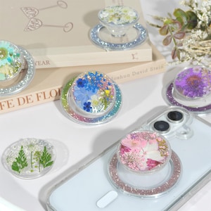May include: Several clear phone grips, each containing dried flowers in shades of blue, pink, and purple. The grips have glittery ring bases in silver, pink, and rainbow hues. Some are attached to a white phone. The grips are a decorative accessory.