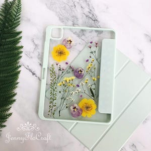 Könnte beinhalten: Eine hellgrüne iPad-Hülle mit einer durchsichtigen Rückseite, die mit gepressten Blumen verziert ist. Die Blumen sind gelb, lila und weiß. Die Hülle hat einen integrierten Ständer.