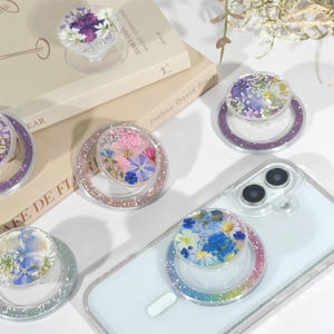 May include: A collection of clear phone grips with embedded dried flowers in various colors. Each grip has a glittery ring around the base. Some grips are attached to a white smartphone.