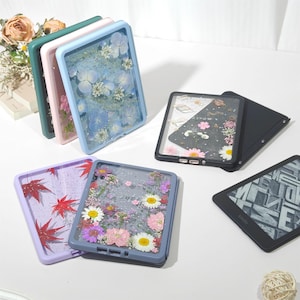 Puede incluir: Una colección de fundas rectangulares para lectores electrónicos con respaldos transparentes incrustados con diversas flores y hojas prensadas. Las fundas vienen en colores como azul claro, rosa, verde oscuro, negro, morado y gris, con un lector negro.