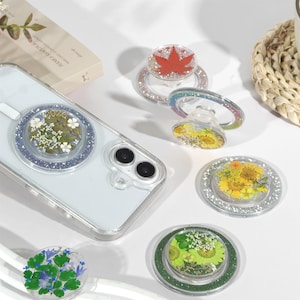 May include: Several clear phone grips with floral and leaf designs, including a red maple leaf and yellow flowers. The grips are decorated with glitter and attached to a clear phone case, showcasing a variety of styles.