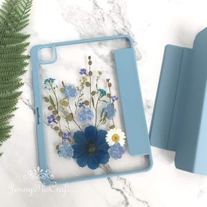 May include: A light blue tablet case featuring a clear back panel decorated with pressed blue and white flowers. The design includes a large dark blue central flower, smaller light blue blossoms, white daisies, and thin green stems and leaves.