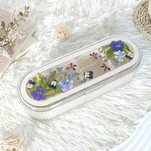 Puede incluir: Una caja blanca rectangular con tapa transparente, adornada con flores secas en tonos azules, morados y blancos. El nombre "Harriet" es visible en el interior. La caja está sobre una superficie blanca y esponjosa.
