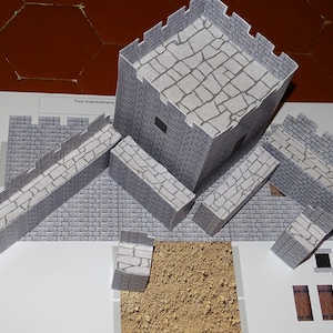 May include: A paper model kit of a medieval castle with gray stone walls and a brown dirt ground. The kit includes a tower, walls, and a walkway. The text "Tour intermediaire" is printed on the paper.