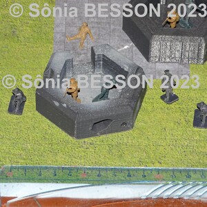 May include: A tabletop wargaming scene with miniature figures and a gray hexagonal bunker. The bunker has a small opening on one side. The figures are in various poses, some holding weapons. The scene is set on a green grassy surface. © Sonia BESSON 2023