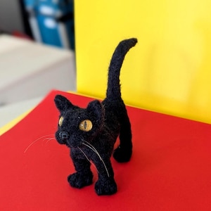 Pode incluir: Uma estatueta de gato preto feita de lã feltrada com uma superfície texturizada, em pé com a cauda levantada. O gato apresenta olhos amarelos proeminentes com pupilas pretas, um pequeno nariz preto e longos bigodes brancos finos.