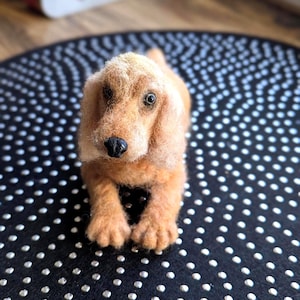 Puede incluir: Figura de perro artesanal de fieltro marrón claro con nariz negra y ojos expresivos. El perro está tumbado sobre una alfombra negra con lunares blancos. La figura está hecha de lana suave y texturizada.