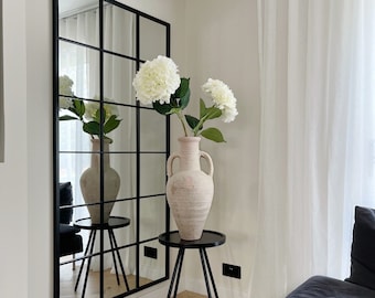 May include: A large black framed mirror with a grid pattern is mounted on a white wall. A black side table with a vase of white flowers sits in front of the mirror. The vase is a light beige color. A black sofa is in the corner.