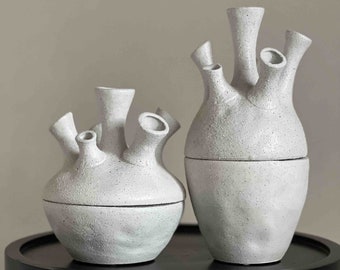 May include: Two white ceramic vases with multiple openings on top. The vases are textured and have a speckled finish. The vases are sitting on a black round table.