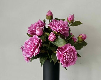 May include: A bouquet of pink peonies in a black cylindrical vase. The vase is sitting on a white linen tablecloth.