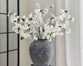 May include: A white faux cherry blossom branch in a gray ceramic vase on a black pedestal.