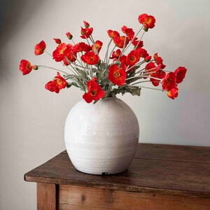May include: A bouquet of bright orange poppy flowers in a white ceramic vase on a wooden table.