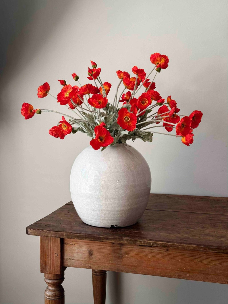 May include: A bouquet of bright orange poppy flowers in a white ceramic vase on a wooden table.