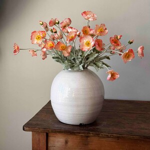 May include: A bouquet of peach-colored poppy flowers in a white ceramic vase on a wooden table.
