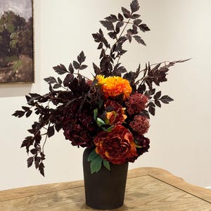 May include: A dark vase displays a colourful floral arrangement. The bouquet includes deep red peonies, orange dahlias, and burgundy foliage. Green leaves accent the base, creating a contrast against the dark vase.